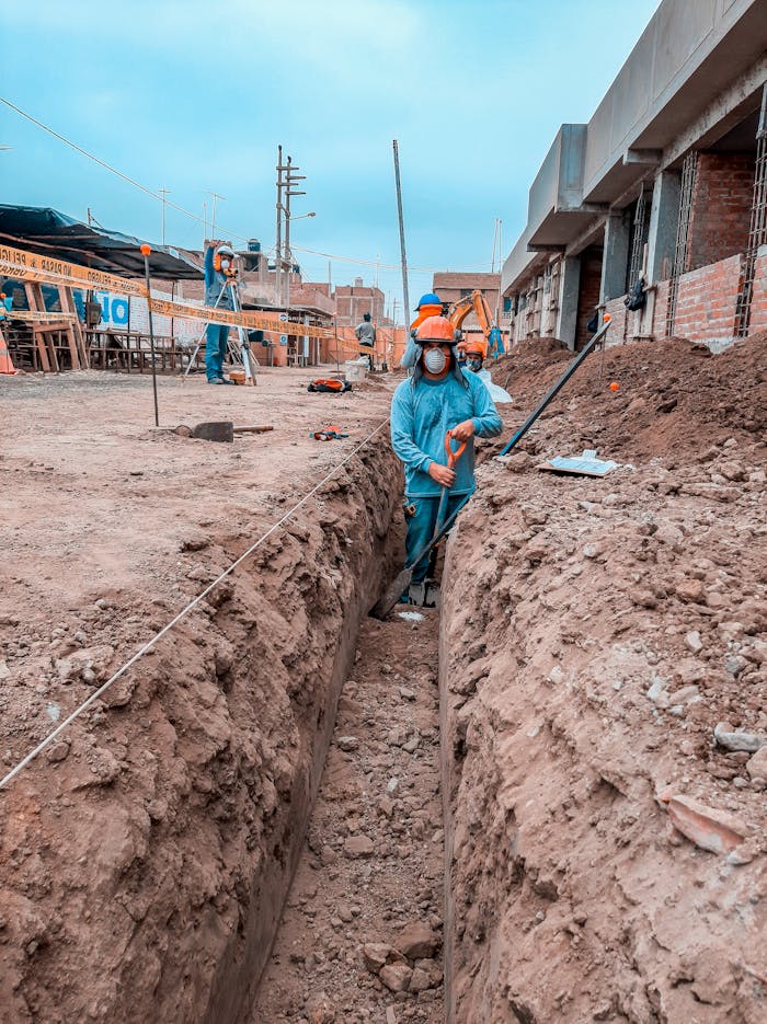 Construction workers in protective gear digging a trench at a site with buildings in the background.