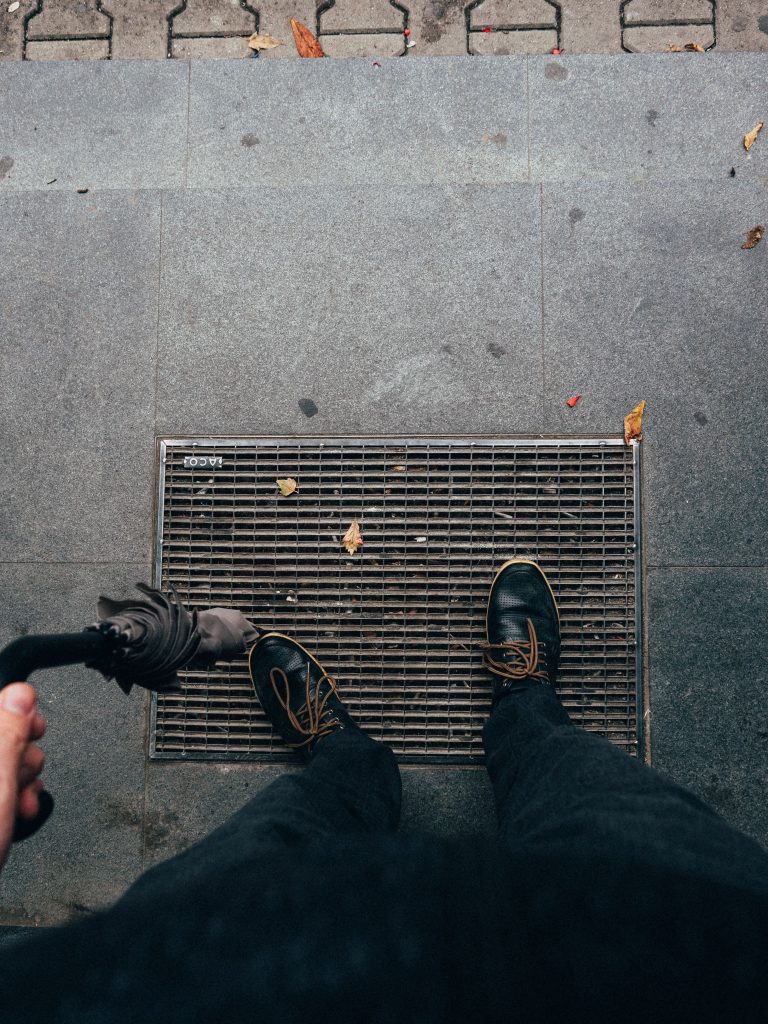 From above of crop anonymous man in dark shoes and black trousers with umbrella standing on ground with fallen leaves and manhole